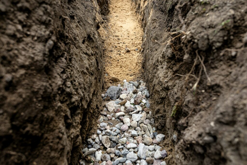 A trench dug in the yard along the fence to lay the drainage pipe,visible gravel and yellow sand.